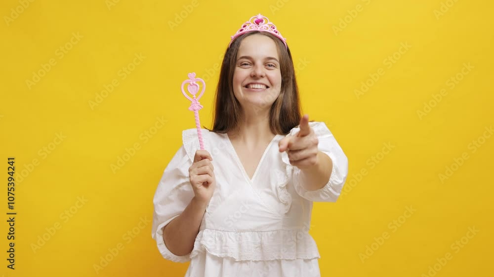 A cheerful woman dressed as a princess holds a wand against a vibrant yellow backdrop A cheerful woman dressed as a princess holds a wand against a vibrant yellow backdrop