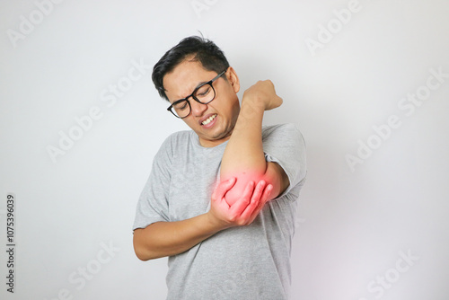 Unhealthy man holds aching elbow. Hitting hand, young man frowns at joint, hisses in pain, tries to alleviate strong pain impulses. Sore bones, articulations. Studio portrait isolated on background.