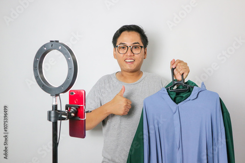 Excited young Asian men is holding up a pair of pants in front of a rack of clothes while showing thumb up gesture. Host live shopping concept.