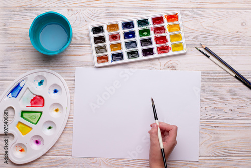 Top View of Child’s Hand Holding Paintbrush with Watercolors on Wooden Background