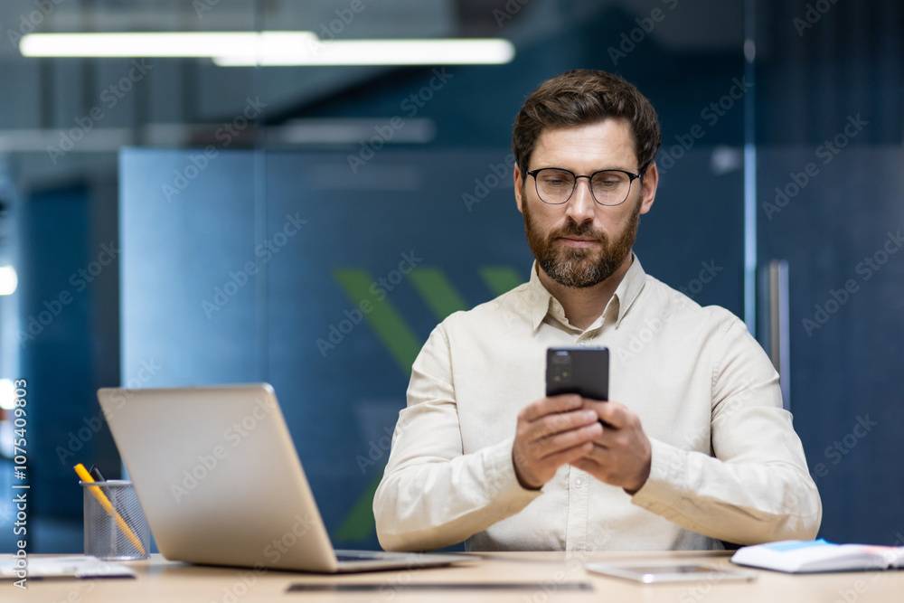 A serious and focused young man is sitting in the office at a desk with a laptop and using a mobile phone A serious and focused young man is sitting in the office at a desk with a laptop and using a mobile phone