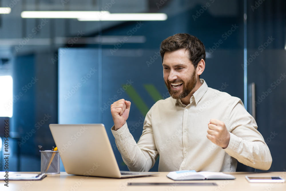 Happy young man of success and result showing yes gesture with hands, sitting in office at desk, looking at laptop screen Happy young man of success and result showing yes gesture with hands, sitting in office at desk, looking at laptop screen