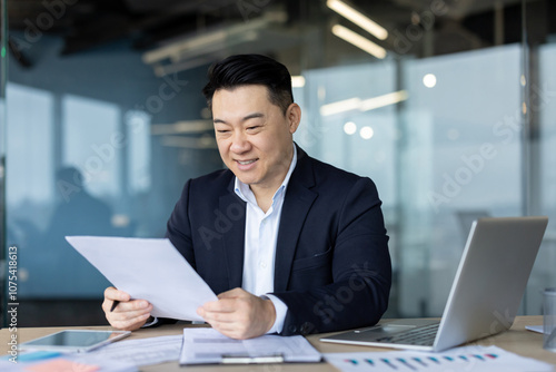 Asian young smiling male businessman sits in the office at a desk with a laptop in a business suit and reads documents in his hands