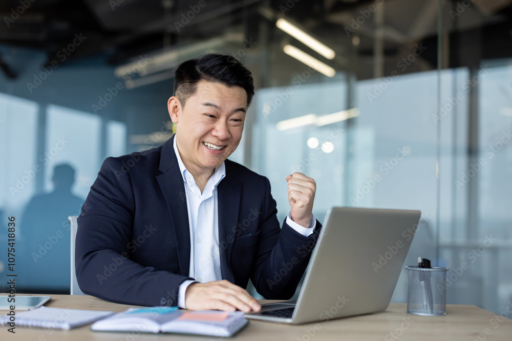 Happy young Asian male businessman sitting in the office at the work desk and being happy, looking at the laptop screen and showing a victory yes gesture with his hand Happy young Asian male businessman sitting in the office at the work desk and being happy, looking at the laptop screen and showing a victory yes gesture with his hand