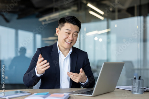 Asian young male businessman sitting in the office at the desk in a suit and talking on a video call on a laptop gesturing with his hands