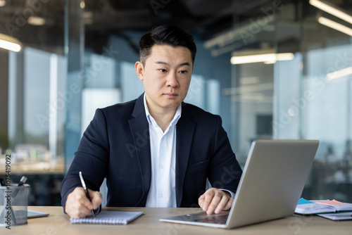 A serious and focused young Asian man in a business suit is sitting in the office at the desk, writing in a notebook and looking at the laptop screen