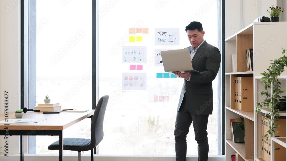 Asian businessman in formal suit discusses graphs and charts during online video call. Standing by panoramic window in modern office, he uses laptop for engaging presentation. Asian businessman in formal suit discusses graphs and charts during online video call. Standing by panoramic window in modern office, he uses laptop for engaging presentation.