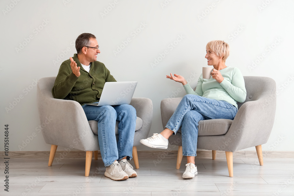 Senior couple with laptop and cup of tea sitting in armchairs and talking near white wall Senior couple with laptop and cup of tea sitting in armchairs and talking near white wall