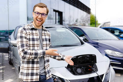 A young man stands next to his new electric car and rests while the car is charging