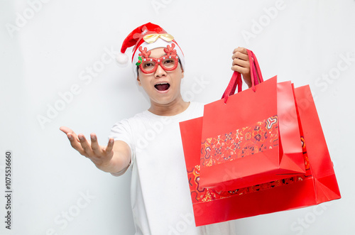 euphoric young asian man wearing santa claus hat and christmas glasses carrying red shopping bag and making yes gesture. christmas sale.