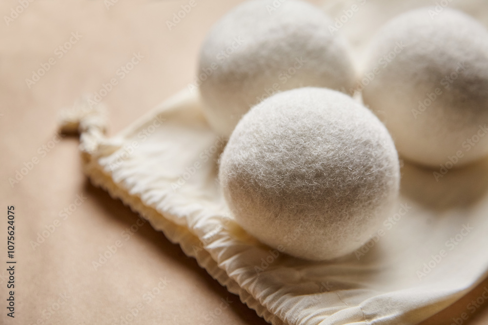 Close-up of three white wool dryer balls resting on an open cotton drawstring bag against a soft brown background, highlighting eco-friendly laundry essentials Close-up of three white wool dryer balls resting on an open cotton drawstring bag against a soft brown background, highlighting eco-friendly laundry essentials