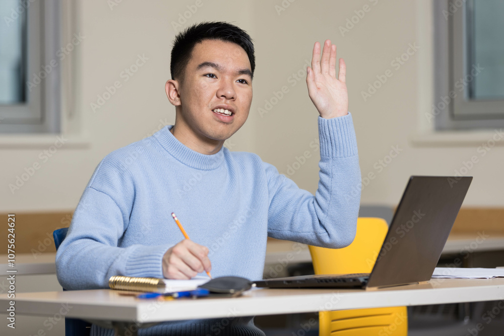 A college student is sitting at a desk raising his hand enthusiastically A college student is sitting at a desk raising his hand enthusiastically