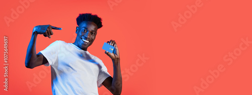 Black man is standing in front of a red backdrop, smiling and holding a credit card in his right hand. He is wearing a white t-shirt and pointing to himself with his left hand.