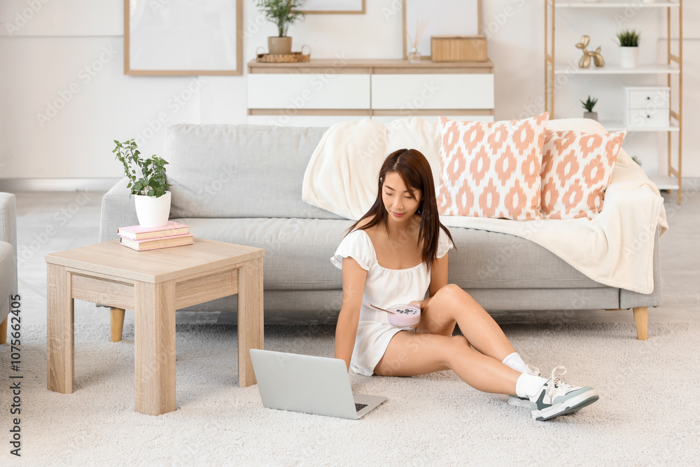 Pretty young Asian woman with laptop eating tasty yogurt at home Pretty young Asian woman with laptop eating tasty yogurt at home