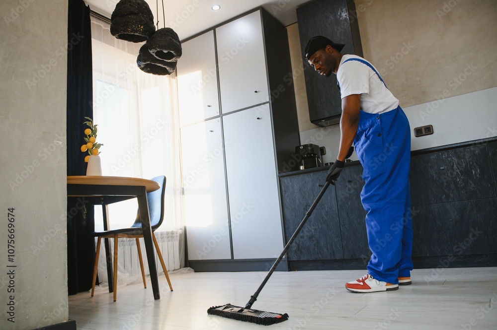 Fototapeta premium Man janitor with mop in hands from cleaning service washing gray floor in uniform.