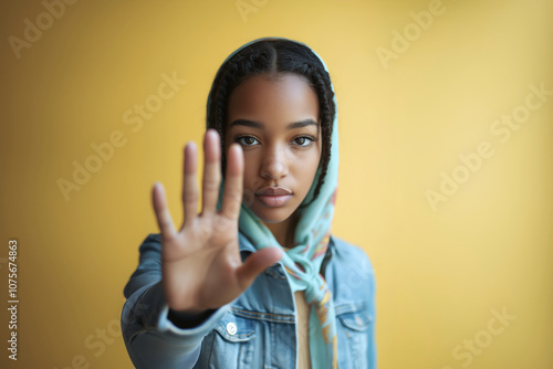 Young woman wearing a headscarf and denim jacket raises her hand in a gesture symbolizing opposition to systemic racism. Her serious expression emphasizes the call for justice and equality. Yellow bac photo