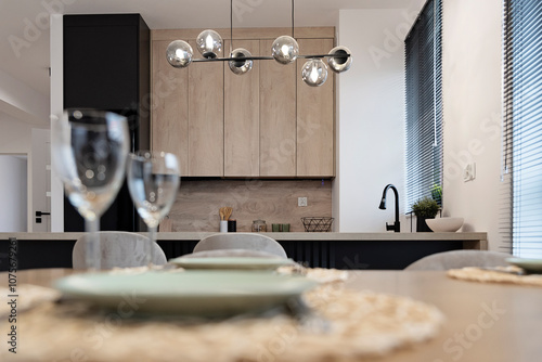 Modern dining room interior with kitchen with wooden counter and dining table with empty plate and glass. Design furniture with stylish glass lapm over the table. Real estate. photo