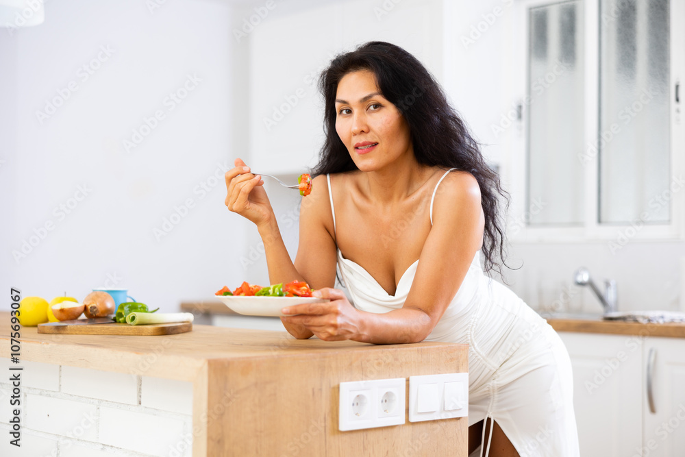 Portrait of asian woman in nightie eating salad in kitchen at home. Woman leaning on kitchen table, eating salad. Portrait of asian woman in nightie eating salad in kitchen at home. Woman leaning on kitchen table, eating salad.
