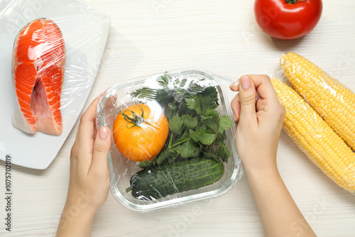 Woman putting plastic food wrap over glass container with vegetables at white wooden table, top view photo