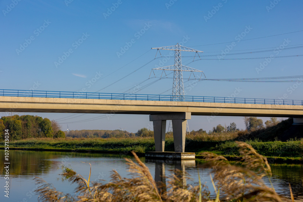 Countryside landscape with road and concrete bridge crossing Lage vaart canal, Electric high voltage pole under blue sky in Autumn, Reed plant and grass along the shore, Almere, Flevoland, Netherlands Countryside landscape with road and concrete bridge crossing Lage vaart canal, Electric high voltage pole under blue sky in Autumn, Reed plant and grass along the shore, Almere, Flevoland, Netherlands