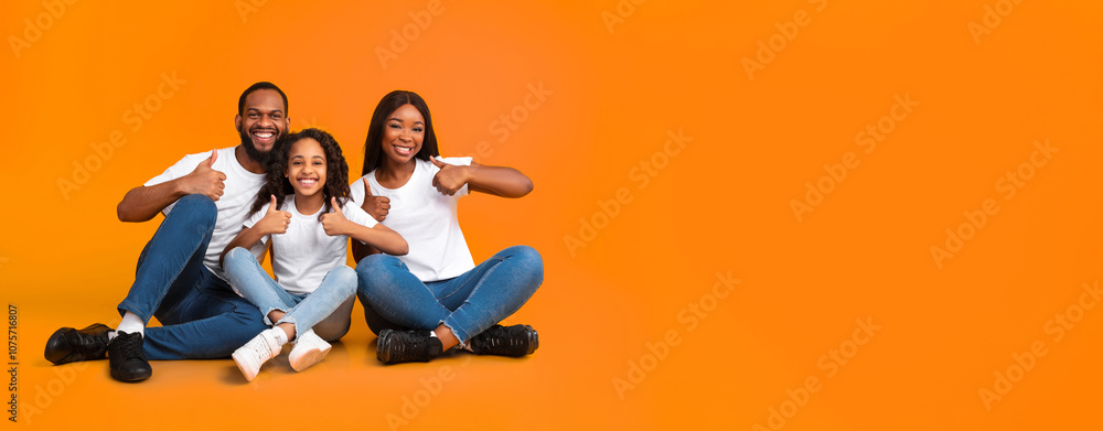 Smiling African American family of three showing thumbs up gesture with both hands, approving or recommending something good, sitting on the floor at yellow studio wall, panorama, free copy space Smiling African American family of three showing thumbs up gesture with both hands, approving or recommending something good, sitting on the floor at yellow studio wall, panorama, free copy space