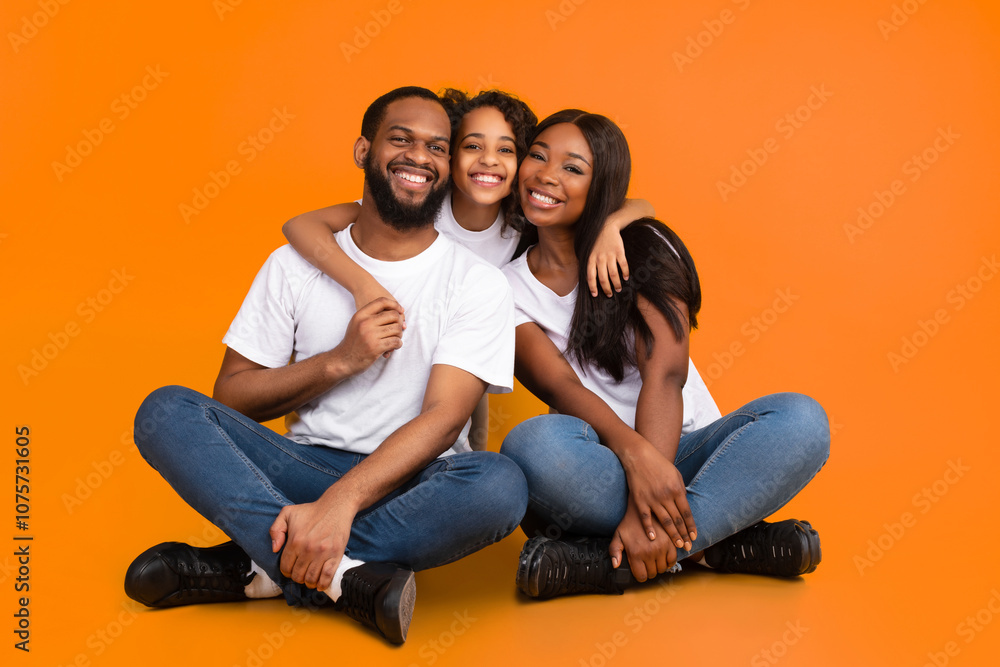 Happy African American man, woman and girl sitting on the floor isolated on yellow studio wall. Smiling daughter emracing her mother and father, looking at camera. Healthy Family Relationship Concept Happy African American man, woman and girl sitting on the floor isolated on yellow studio wall. Smiling daughter emracing her mother and father, looking at camera. Healthy Family Relationship Concept