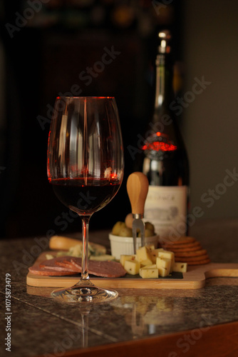 Glass of a red whine on a kitchen island, cheese, olives and creckers catted and placed on a wooden cheese board, bottle of red wine as a background. Dark colors and luxury lifestyle photo