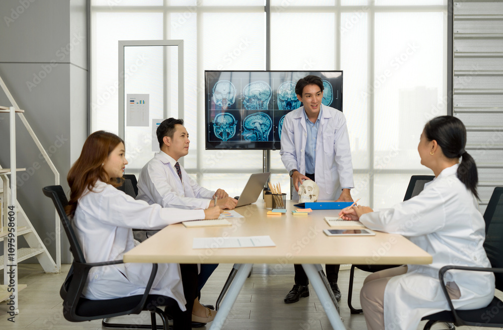Young medical professional standing and presenting, holding a model of human skull and referencing image of brain scan displayed on a screen. The audience listening attentively with the presentation. Young medical professional standing and presenting, holding a model of human skull and referencing image of brain scan displayed on a screen. The audience listening attentively with the presentation.
