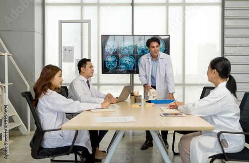 Young medical professional standing and presenting, holding a model of human skull and referencing image of brain scan displayed on a screen. The audience listening attentively with the presentation.