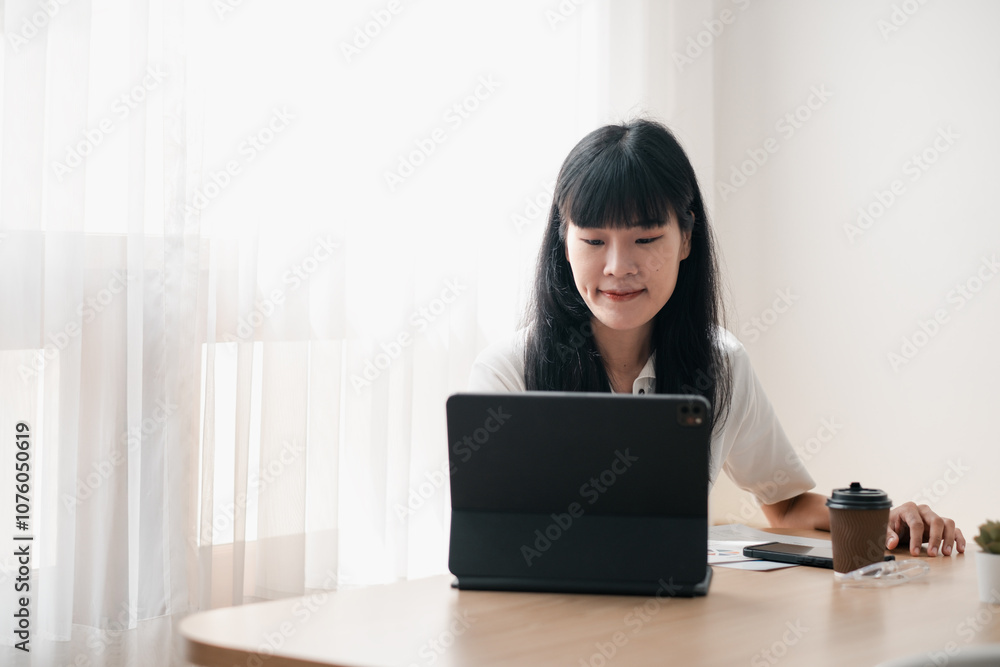 A young woman focused on her tablet in a home office setting, with coffee and documents nearby, illuminated by natural light. A young woman focused on her tablet in a home office setting, with coffee and documents nearby, illuminated by natural light.