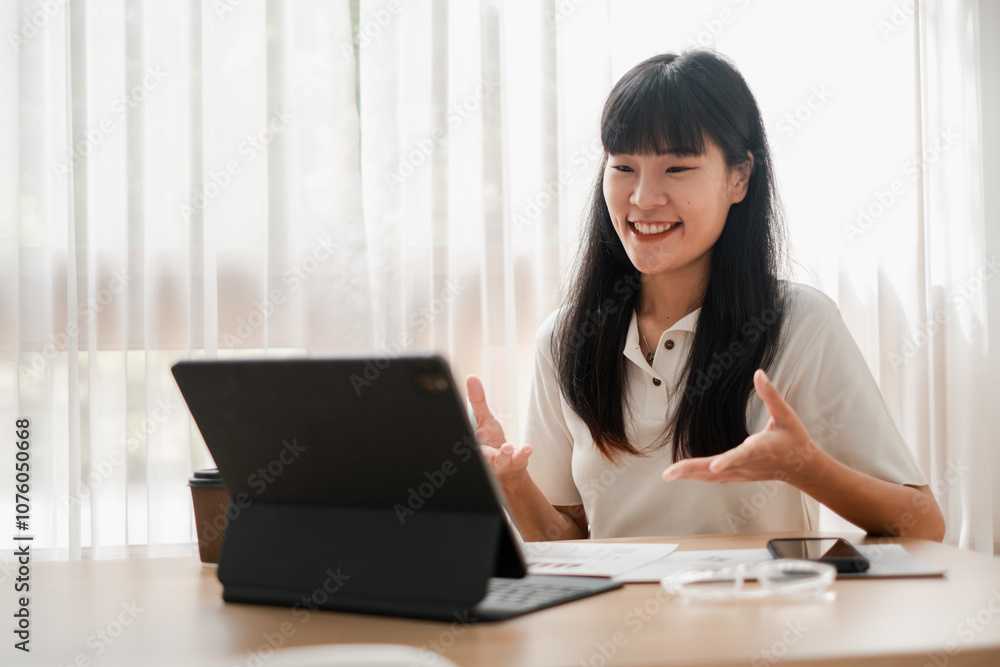 A cheerful woman in a white shirt having a video call on a tablet, sitting at a desk with natural light filtering through curtains. A cheerful woman in a white shirt having a video call on a tablet, sitting at a desk with natural light filtering through curtains.