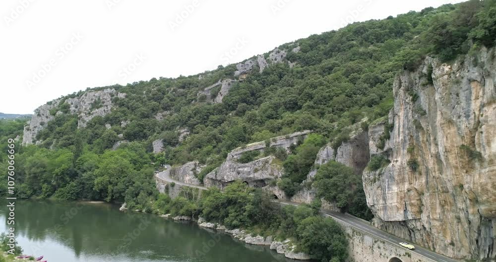 Aerial view of Ardèche, France, with a drone flying backward as two cars pass by on a winding road near a river and rocky cliffs Aerial view of Ardèche, France, with a drone flying backward as two cars pass by on a winding road near a river and rocky cliffs