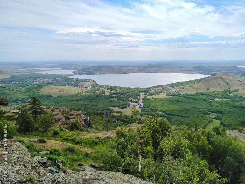 A breathtakingly stunning view of a serene and peaceful lake from the heights of a towering mountain, surrounded by vibrant lush greenery and a beautiful sky filled with fluffy white clouds A breathtakingly stunning view of a serene and peaceful lake from the heights of a towering mountain, surrounded by vibrant lush greenery and a beautiful sky filled with fluffy white clouds