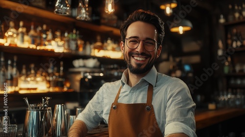 Happy male bartender smiling warmly, wearing glasses and an apron, in a cozy bar with warm lighting and shelves of bottles.