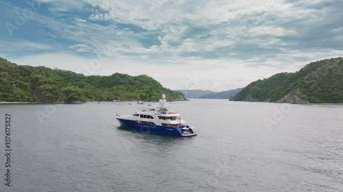 An aerial view captures a luxury yacht gliding over the tranquil waters of Puntarenas, Costa Rica, framed by green hills and a cloudstudded sky, creating a breathtaking scene photo