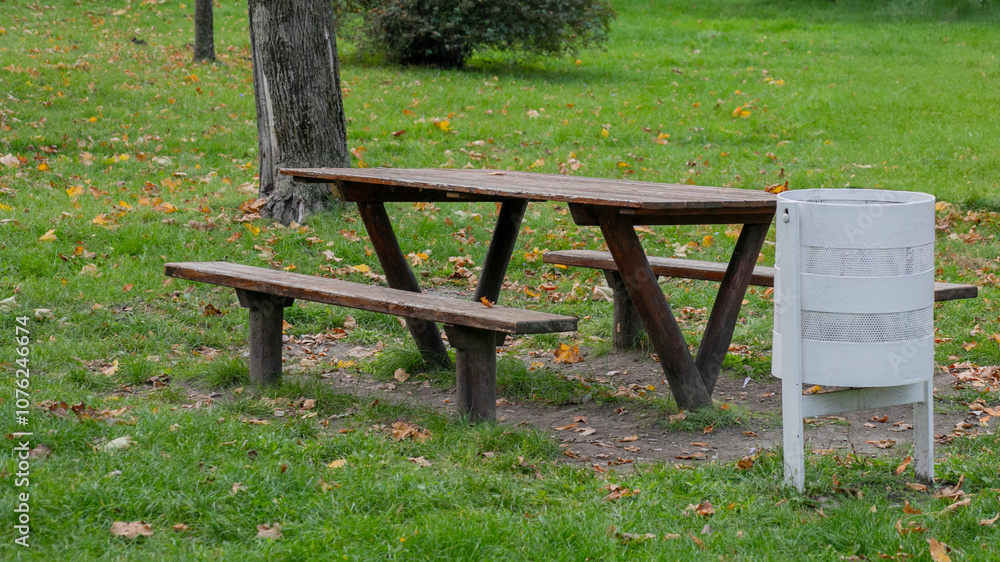 Wooden picnic table with benches and a white trash can on green grass, related to environmental awareness and outdoor gatherings Wooden picnic table with benches and a white trash can on green grass, related to environmental awareness and outdoor gatherings