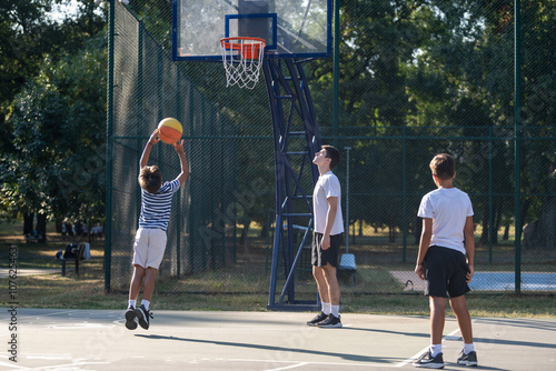 Boys engage in outdoor basketball training with a coach, practicing dribbling, passing, and shooting skills on a court surrounded by greenery under bright natural light, fostering teamwork