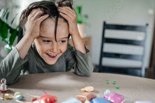 Cheerful boy with head in hands sitting near clay at table photo