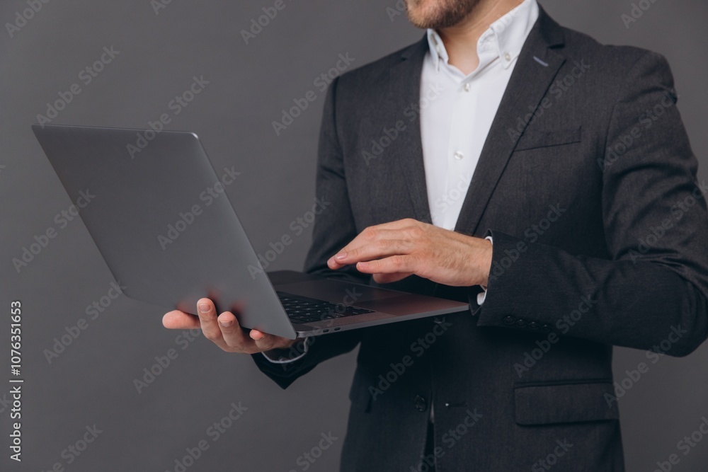 Close-up portrait of successful and confident businessman in grey suit, working with laptop, standing over gray background Close-up portrait of successful and confident businessman in grey suit, working with laptop, standing over gray background