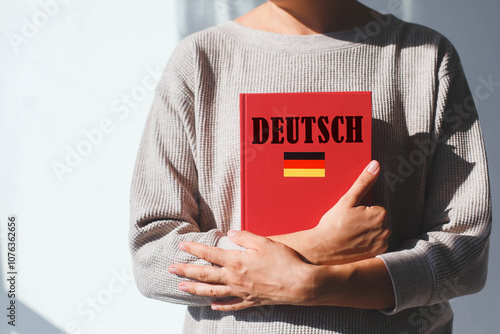 Photography young woman in beige sweater holds red german language textbook with german inscription and german flag, foreign language learning concept