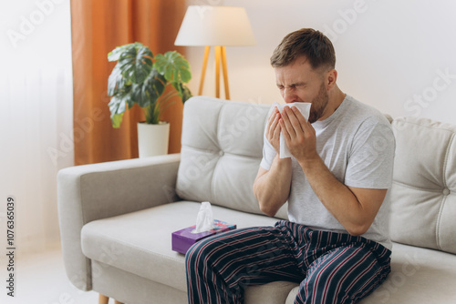 Fotografia Unhappy middle aged man suffering from fever and flu, blowing nose in napkin, sitting in living room interior