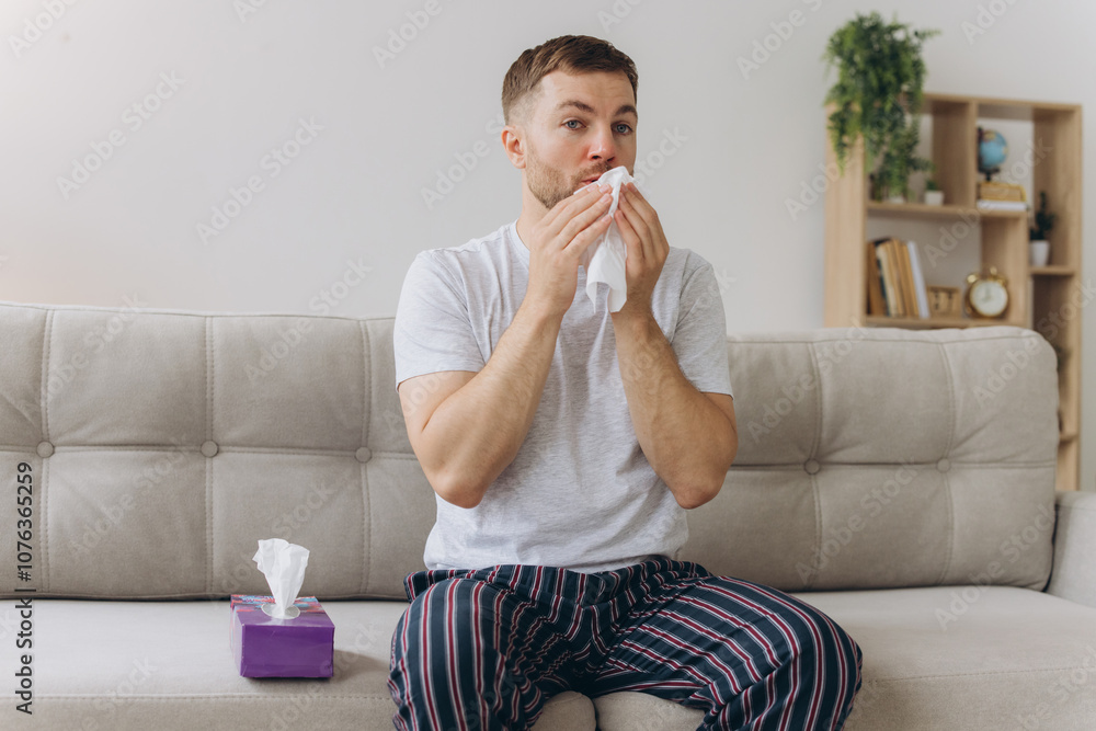 Unhappy middle aged man suffering from fever and flu, blowing nose in napkin, sitting in living room interior. Covid-19 lockdown, treatment of illness, cold and runny Unhappy middle aged man suffering from fever and flu, blowing nose in napkin, sitting in living room interior. Covid-19 lockdown, treatment of illness, cold and runny