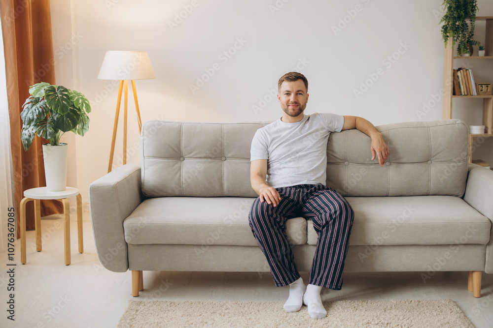 Joyful young bearded man in pajamas sitting on a sofa in living room at home with copy space Joyful young bearded man in pajamas sitting on a sofa in living room at home with copy space
