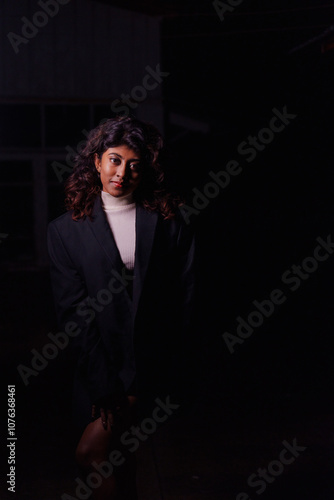 A woman in a black suit and white sweater stands in a dark room. She is smiling and looking at the camera