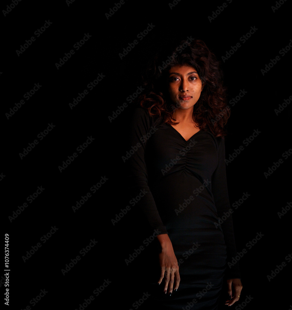 A woman in a black dress stands in front of a dark background. She has a serious expression on her face A woman in a black dress stands in front of a dark background. She has a serious expression on her face