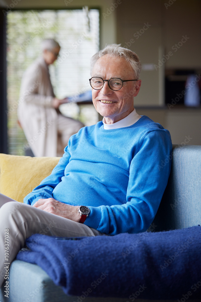 Portrait of smiling senior man sitting on couch at home with woman in background Portrait of smiling senior man sitting on couch at home with woman in background
