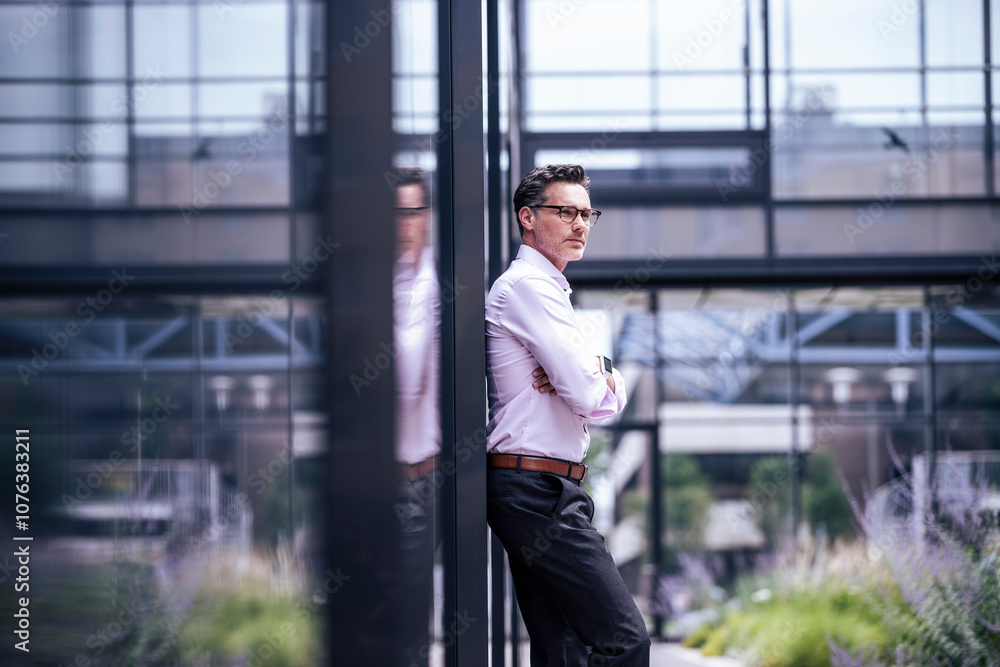 Thoughtful businessman with arms crossed leaning on glass wall Thoughtful businessman with arms crossed leaning on glass wall