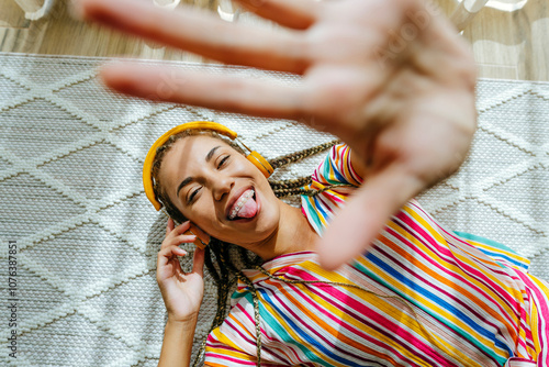 Cheerful young woman wearing wireless headphones and showing stop gesture on carpet at home photo