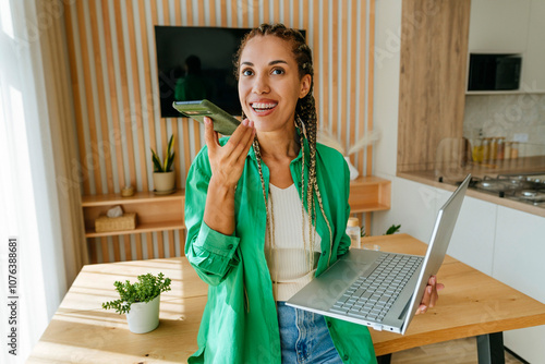 Happy freelancer sending voicemail through smart phone and standing with laptop at home photo