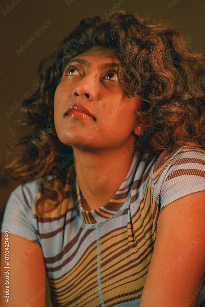 A woman with brown hair and a blue and yellow striped shirt is looking up. She has a red lip and a necklace